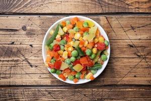 Chopped frozen vegetables in a plate on a wooden background. Corn peas pepper carrots. copy space. photo