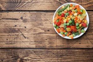 Chopped frozen vegetables in a plate on a wooden background. Corn peas pepper carrots. copy space. photo