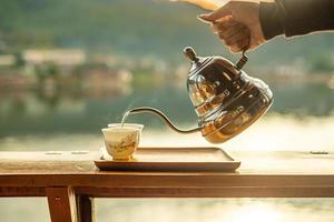 Hand holding vintage teapot and pouring hot tea to cup on wood table against lake view background at coffee shop in the morning sunrise, Ban Rak Thai village, Mae Hong Son province, Thailand photo