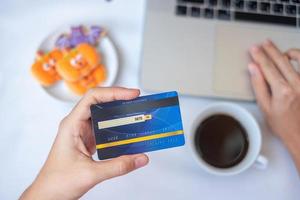 mano que sostiene la tarjeta de crédito para compras en línea en la computadora portátil mientras come galletas de halloween y café. feliz halloween, hola octubre, otoño otoño, festivo, fiesta y concepto de vacaciones foto