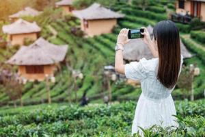 mujer turista feliz con vestido blanco tomando fotos con un teléfono inteligente móvil en un hermoso jardín de té. viajero visitando el pueblo tailandés de ban rak, mae hong son, tailandia. concepto de viajes, vacaciones y vacaciones