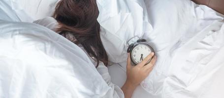 alarm clock and asian woman hand stop time in bed while sleeping, young adult female wake up late in the morning. Relaxing , sleepy, daily routine and have a nice day concepts photo