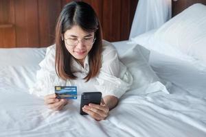 woman using mobile smart phone and credit card for online shopping while making order on bed in morning at home. technology, ecommerce, digital banking online payment and apartment living concept photo