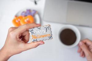 Hand holding Halloween Cookie and drinking coffee during using computer laptop. Happy Halloween, online shopping, Hello October, fall autumn, Festive, party and holiday concept photo