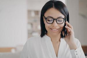 Smiling businesswoman wearing glasses talking on smartphone with client, answering call in office photo