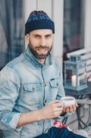 Vertical shot of attractive blue eyed bearded male in stylish clothing, drinks hot tea, looks with pleased expression at camera, spends recreation time at terrace cafe, smiles gently at camera photo