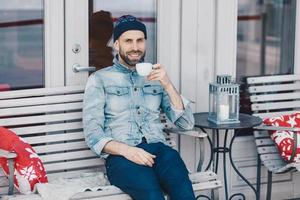 feliz hombre de ojos azules con aspecto positivo, se sienta en el interior acogedor de la cafetería de la terraza, disfruta del café aromático, tiene tiempo libre después del trabajo. el hombre guapo tiene barba, mira alegremente a la cámara. foto