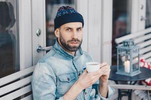Close up shot of handsome serious bearded male, looks directly into camera with blue eyes and confident expression, drinks hot beverage, enjoys spare time. People, drinks, lifestyle concept. photo