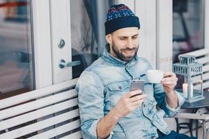 Delighted happy male with thick beard and mustache, reads positive news on cell phone, enjoys recreation time, drinks tea in coffee shop, has happy expression. Smiling handsome man uses cellular photo