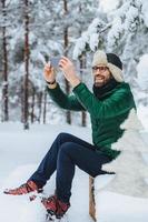 el retrato vertical de un hombre de aspecto agradable hace fotos con un teléfono inteligente, toma fotos de hermosos paisajes invernales, disfruta de la naturaleza y el clima helado, tiene una sonrisa agradable. personas, concepto de tecnología