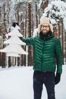 el retrato de un alegre y apuesto hombre barbudo lleva un sombrero caliente, un anorak verde y guantes sostiene un pequeño abeto artificial blanco, se encuentra en un bosque invernal cubierto de nieve. hermosos paisajes foto