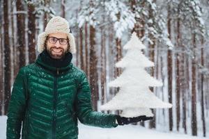 personas, emociones, clima nevado y concepto de temporada. feliz y alegre joven sin afeitar vestido con ropa de abrigo, sostiene un abeto, se para contra el fondo de los árboles, disfruta de hermosos paisajes naturales foto
