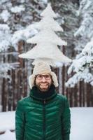 Vertical portrait of cheerful bearded man has fun alone in winter forest, keeps artificial fir tree, poses outdoors, admires frosty snowy weather, expresses positiveness and pleasant emotions photo