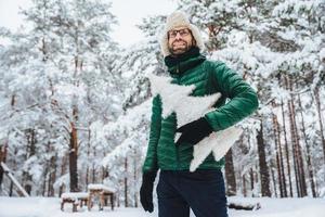 alegre modelo masculino vestido con ropa de invierno, sostiene un abeto artificial blanco, se para en el bosque de invierno, tiene una sonrisa agradable, disfruta de hermosos paisajes. gente, vacaciones, concepto de recreación foto