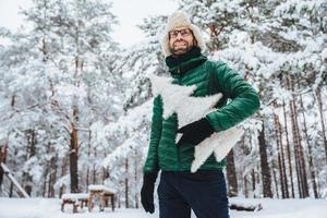 Cheerful male model dressed in winter clothes, holds white artificial fir tree, stands in winter forest, has pleasant smile, enjoys beautiful landscapes. People, holidays, recreation concept photo