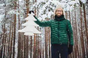 apuesto hombre sin afeitar con anteojos, usa ropa abrigada de invierno, sostiene un abeto blanco, tiene buen humor, anticipa el año nuevo y la navidad, posa contra los árboles altos de invierno foto