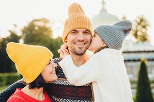 Pleasant looking little girl being thankful to her father for buying new bicycle, kisses him in cheek. Young family walk together outdoor, spend spare time in park, wear knitted fashionable hats photo