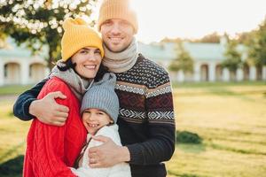 Horizontal portrait of young handsome man in yellow hat and warm knitted sweater, embrace his wife and daughter, pose at camera while stand in park. Three family members spend weekends together photo