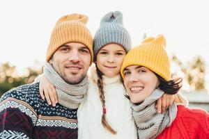 Portrait of adorable small girl wears knitted hat and sweater stands between parents, embrace them. Beautiful woman wears warm scarf and sweater enjoys free time with husband and little daughter photo