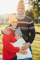 Vertical portrait of friendly family couple stand together, embrace each other, have good relationships, enjoy sunny weather. Handsome unshaven man hugs his wife and daughter, pose outdoors photo