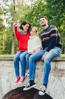 una mujer tranquila y despreocupada, un hombre y una niña pequeña y linda se sientan en el puente al aire libre, admiran el amanecer. madre cariñosa le muestra a su pequeña hija la belleza de la naturaleza. la familia disfruta pasar el tiempo libre en el parque foto