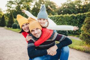 Handsome young man spends his free time with family, recieves embrace from adorable daughter and pretty wife, have picnic during autumn weekend. Happy affectionate family have good relationships. photo