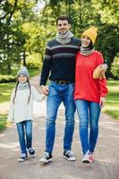 Portrait of friendly family walk across park. Beautiful brunette female in knitted yellow hat and sweater, handsome man embraces his wife and holds daughters hand, enjoy autumn weather and fresh air photo