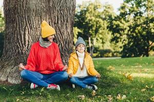 Body language concept. Carefree little kid sits near her beautiful mother, keep hands in mudra sign, keep eyes closed, try to calm down after hard work at home, admire fresh air in beautiful park photo