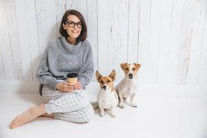 Beautiful brunette woman sits on floor with her two favourite dogs, dressed casually, drinks takeaway coffee. Pleased female enjoys calm domestic atmosphere. People and animals, good relations photo