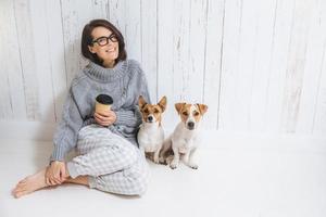 Smiling cheerful young female wears warm woolen sweater, square eyewear, drinks hot beverage, relaxes on floor and her two favourite pets, look directly into camera. People and animals conecpt photo