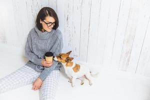 People, lifestyle and leisure concept. Carefree beautiful young woman dressed in domestic clothes, drinks takeaway coffee, spends free time with dog, enjoys calm atmosphere, being in good mood photo