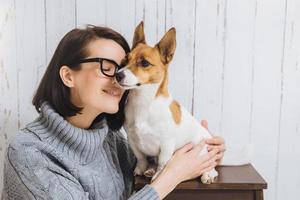 Shot of attractive young woman embraces her favourite dog, touches with nose, expresses great love to pet. Loyal dog has good relationships with host. Friendship, relations, animals and people photo