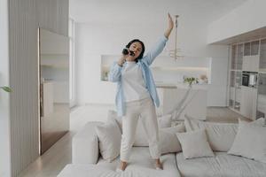 Playful young woman is listening music in earphones, singing in microphone, dancing on sofa. photo