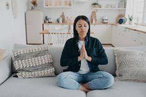 una joven de raza mixta está practicando yoga, sentada en un sofá en posición de loto con los ojos cerrados. foto