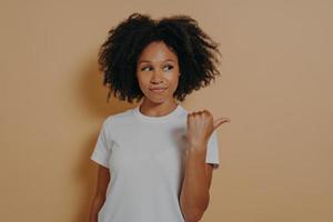 Pretty curly haired african woman points thumb aside, recommends promo and advertises product photo