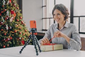 Joyful lady sitting at table with Xmas tree on background in front of phone on tripod photo