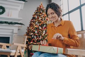 Happy European woman unpacking Xmas gifts while sitting in room with decorated New Year tree photo