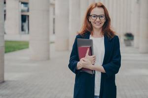 Young confident beautiful ginger woman holding laptop and note book posing outdoors photo
