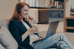 Young dispatcher lady working remotely via wireless headset and laptop, sitting on sofa in teahouse photo