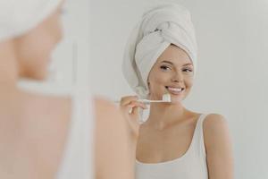 Beautiful young woman with healthy perfect smile brushing teeth and looking in mirror photo