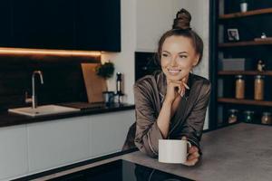 Relaxed positive young woman in silk cozy pajama having cup of coffee in stylish kitchen interior