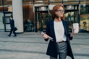 Beautiful redhead female entrepreneur standing outdoors with coffee cup and laptop in hands photo