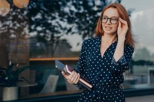 Female entrepreneur carries tablet smartphone and organizer poses near window of restaurant photo