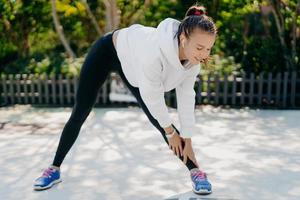 Motivated young adult woman goes in for sport regularly reaches hand to foot leans down has feet shoulder width apart wears sportsclothes enjoys training outdoor sports activity for weight loss photo