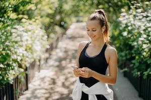 Sport as style of life. Horizontal shot of healthy slim sporty woman focused at smartphone checks information and chooses song from playlist dressed in sportwear poses outdoor breathes fresh air photo