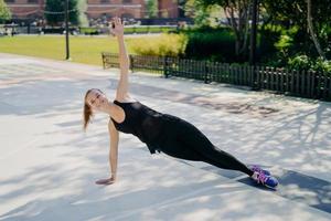 Sport and recreation concept. Athletic slim woman stands in sideways plank raises arm smiles positively dressed in active wear leads healthy lifestyle strengthens core muscles improves stability photo