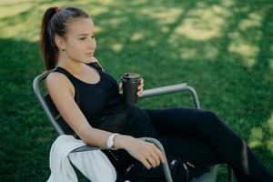 Outdoor shot of thoughtful dark haired beautiful female dressed in sports clothes drinks takeaway coffee spends free time in open air poses on chair at garden enjoys good weather breathes fresh air photo