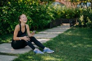 Overjoyed sportswoman sits on path laughs out uses smartphone for listening music from playlist share content and chatting dressed in active wear enjoys spare time. Active lifestyle concept. photo