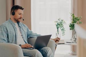 Male freelancer listens attentively tutorials how to start own business writes down information wears stereo headphones and laptop computer works online from home makes video call watches webinar photo