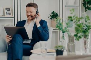 Freelance workday. Cheerful busy man manager works distantly from home online makes video call via laptop computer and headphones discuss working issues with business partner analyzes statistics photo